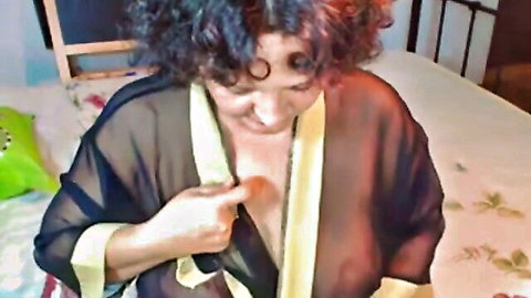 Mature woman in sheer black robe teasing cleavage on bed