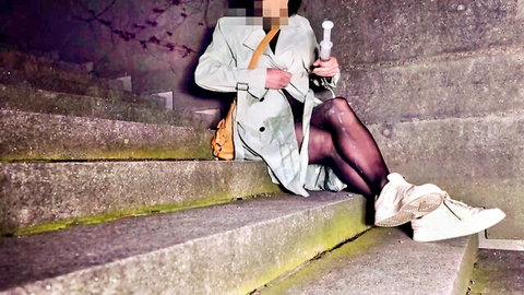 Crossdresser in open trenchcoat and black stockings holding dildo on stairs