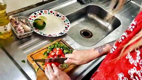 Tattooed woman in red dress chopping tomatoes in kitchen, teasing before sex
