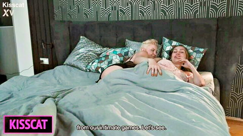 Busty blonde MILF kissing brunette with big tits exposed in lesbian bedroom embrace