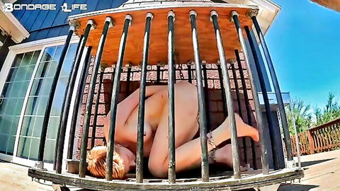 Nude blonde woman in outdoor bondage cage, submissive pose
