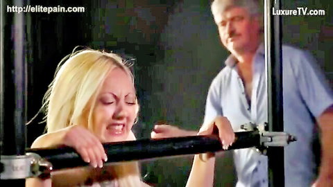 Blonde woman grimacing in pain, restrained for caning in Elite Pain BDSM scene
