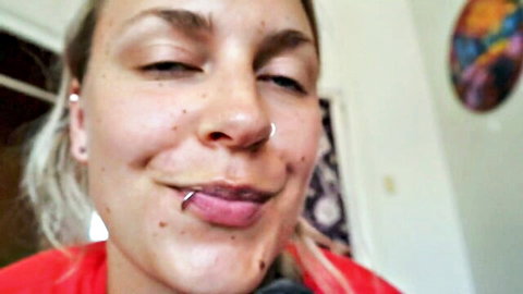 Blonde woman with nose and lip piercings smiling playfully, tongue out