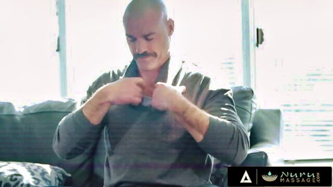 Bald muscular man with mustache sits on couch in gray hoodie during nuru massage