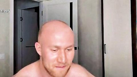 Shirtless bald muscular young hunk with beard in bedroom gazing down