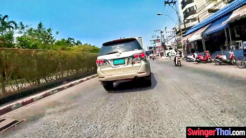 Beige SUV driving uphill on sunny Thai street past motorbikes shops and palms.