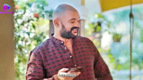 Bald bearded Indian man in unbuttoned maroon shirt holds phone outdoors