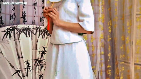 Abby Kitty in elegant white harem dress, holding red tassel, seductive pose