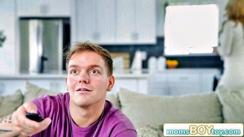 Shocked stepson on couch holds remote staring at busty blonde MILF stepmom in kitchen