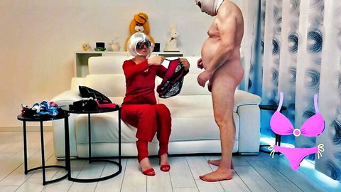 Dominant woman in red catsuit showing lingerie to naked overweight cuckold in feminization scene