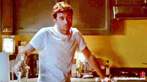 Clothed handsome ginger man in white t-shirt holding bottle in yellow kitchen BDSM scene