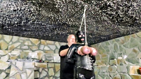Bald latex dom man squeezes exposed pink tits of hooded bound latex slave suspended arms-up in BDSM dungeon.