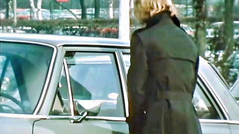 Blonde in trench coat stands by open door of vintage car in parking lot