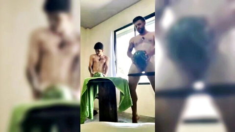 Muscular bearded man stands nude holding watermelon over his cock while shirtless friend leans on table