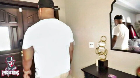 Black man in white tee and cap stands at door in room with PIMPIN logo tattoo
