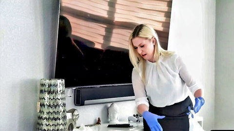 Blonde woman in sheer white blouse black skirt blue gloves bending over desk teasing