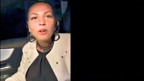 Bald Asian woman in white blazer with hoop earrings opens mouth in car