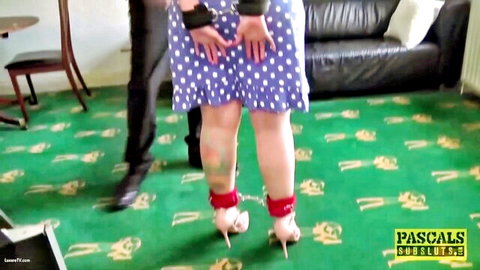 Bound submissive woman in short polka-dot dress with hands cuffed behind back and ankles roped in red straps wearing high heels