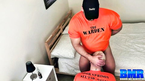 Muscular daddy warden in orange shirt grabs submissive hairy guy's face for rough gay facefuck on bed