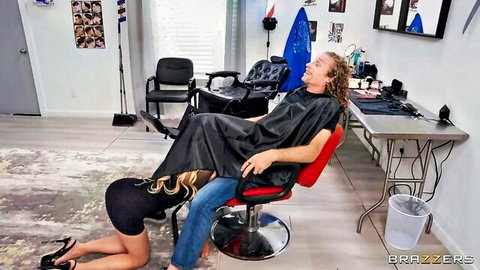 Blonde woman on knees giving blowjob under cape to man in barber chair