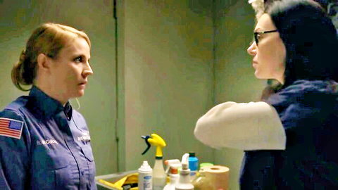 Two uniformed nurses talking intently across table with cleaning supplies in medical room