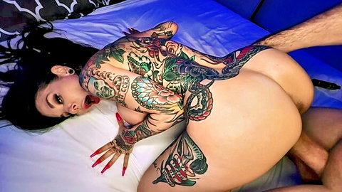 Heavily tattooed brunette with tongue out takes big cock against wet pussy in doggystyle on bed