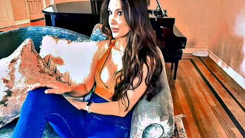 Brunette MILF with big tits lounging seductively in tight orange top and blue pants on luxury sofa