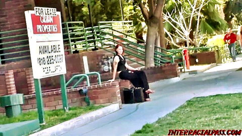 Sexy redhead MILF in black outfit sitting on suitcase by for lease sign outdoors