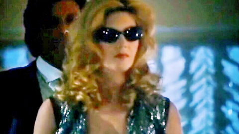 Blonde wife in sparkling green dress and sunglasses enters swing club with suited husband