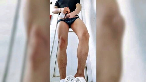 East Asian teen boy sitting legs spread pulling down black shorts exposing hard cock in bathroom