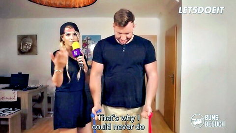 Blonde in short black dress holds yellow mic gesturing at muscular guy in black tee and shorts