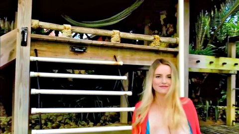 Blonde babe with huge natural tits in skimpy orange bikini smiling in tropical hut