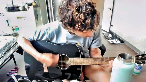 Curly-haired guy in blue t-shirt strumming black acoustic guitar on kitchen floor