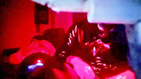 Crystalbitchx in shiny black latex catsuit, seductive pose in red-lit room