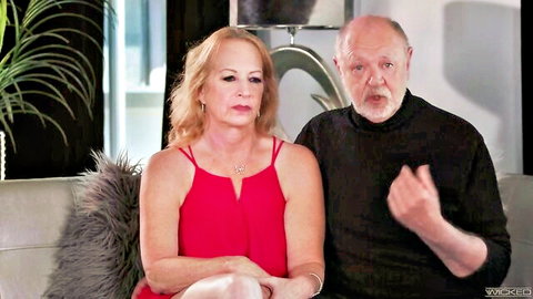 Chubby mature blonde granny in red tank top sits with bald older man on bedroom couch