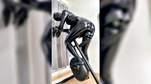 Woman in shiny black latex catsuit bending over on exercise bike, latex fitness fetish pose