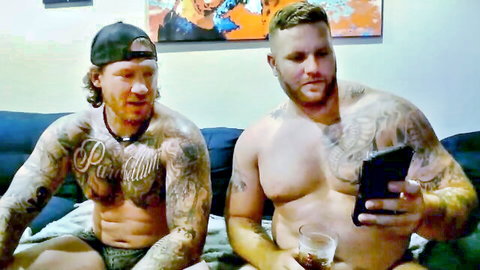 Two muscular tattooed bearded bears lounging shirtless in underwear on couch holding tablet