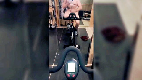 Nude man masturbating on stationary bike in gym workout