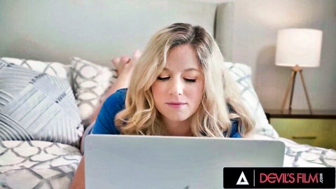 Blonde River Lynn lounges on bed with laptop in tight blue top, teasing viewers