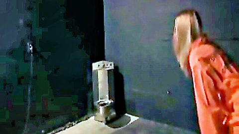 Blonde in shiny orange latex suit standing in dark basement near metal prison toilet