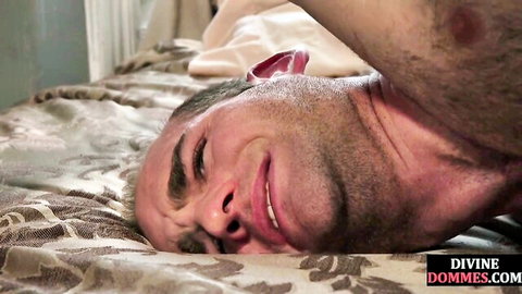 Hairy submissive man grins upside down with head hanging off bed in femdom scene