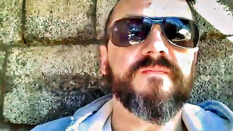 Bald bearded man in sunglasses and hoodie leaning against stone wall