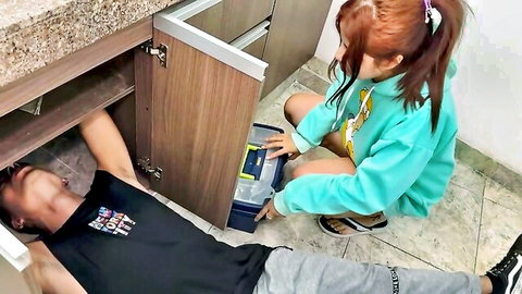 Cute redhead teen in cyan hoodie squats helping guy fix sink under kitchen cabinet