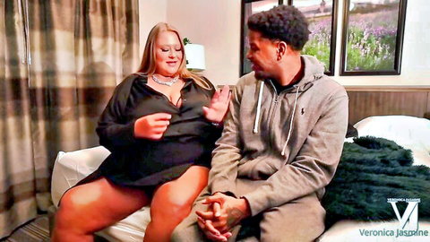 Blonde BBW Veronica Jasmine teases deep cleavage and thick thighs sitting intimately with black stud on bed
