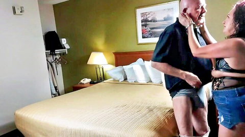 Pink-haired MILF in lacy lingerie top and shorts kisses bald mature man's face in hotel room foreplay