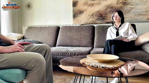 Young Asian therapist in blouse and skirt sits with tattooed male client on office couch