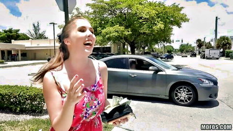 Excited brunette in short pink dress laughs while flashing cash in sunny public parking lot