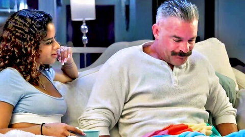 Young curly-haired beauty in tight blue tank top flirts intimately with older mustached man on couch