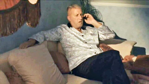Mature man in open shirt sits relaxed on sofa talking on phone