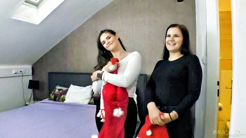 Two smiling brunette girls in sweaters holding red Christmas stockings teasing in bedroom